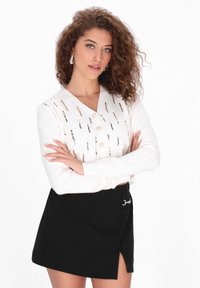 Fehér, gombos kardigán arany és fekete díszítéssel, párosítva egy magas derekú fekete szoknyával, amelynek átlós szabása és fém részletei vannak.