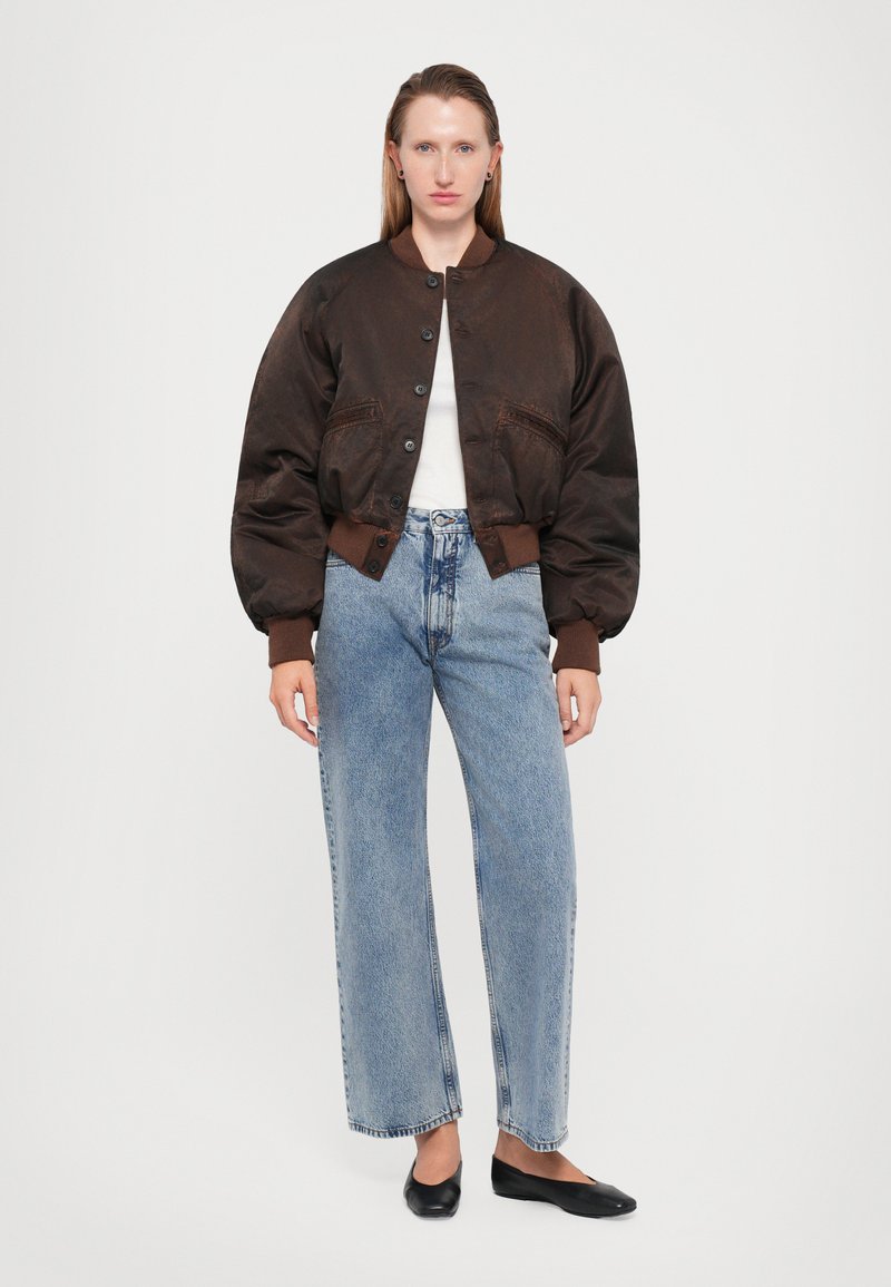 Veste aviateur marron avec un col rond, des boutons et des manches surdimensionnées. Associée à un jean taille haute bleu clair et des ballerines noires.