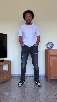 Camisa branca de manga curta, jeans escuros rasgados e ténis pretos. O fundo apresenta um armário de madeira e uma televisão sobre um chão de azulejos.