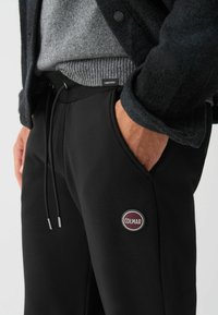 Mustad mustad pusjad, millel on elastne vöökoht ja pael, küljetasku ning ümar punase ja valge "COLMAR" logo reie piirkonnas.
