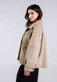 Beige, texturerad ulljacka med krage, framknappar och vida ärmar, i kombination med mörka byxor.