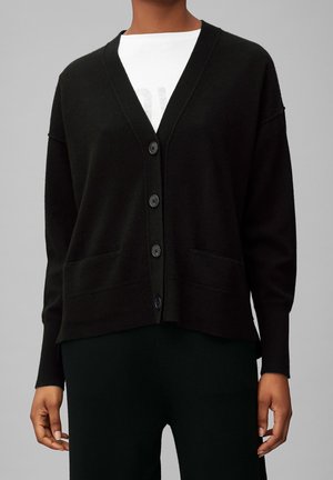 Sort knapper-op cardigan med lange ærmer over en hvid skjorte, båret af en person, der står mod en ensfarvet grå baggrund.