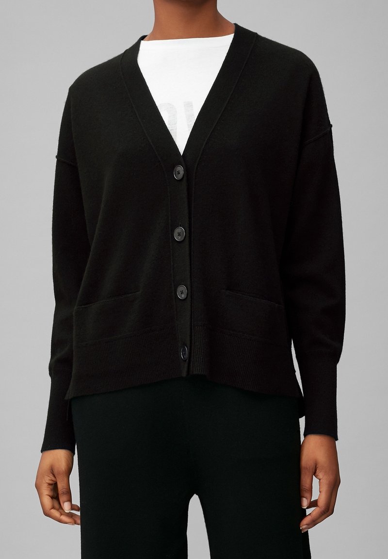 Cardigan noir boutonné avec manches longues porté sur une chemise blanche, porté par une personne debout devant un fond gris uni.