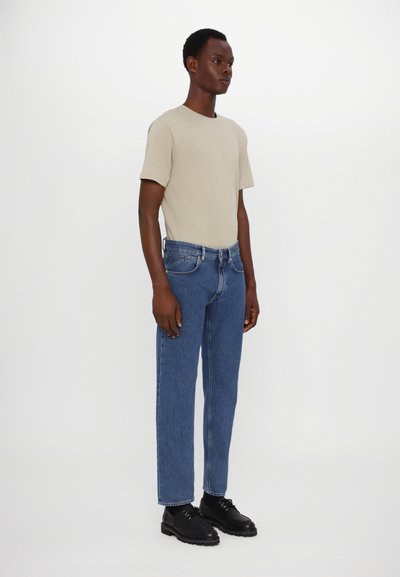 Camiseta de algodón beige, jeans de denim azul de corte recto y zapatos negros de cordones. El conjunto presenta líneas simples y limpias con un detalle mínimo.