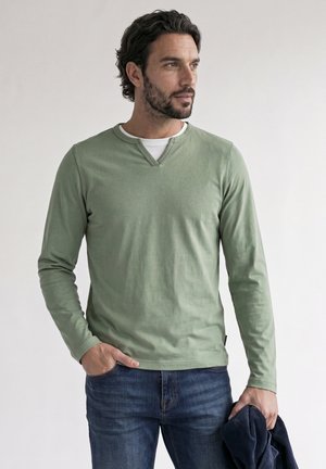 Hombre con cabello rizado y oscuro y barba, vestido con una camisa verde de manga larga y vaqueros azules, sosteniendo una chaqueta oscura en su mano izquierda.