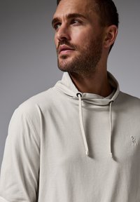 Sudadera con capucha beige, con cordón, textura suave y detalle de logo en el lado izquierdo del pecho. El diseño presenta un escote relajado.