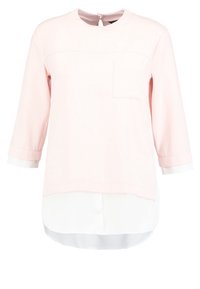 Pull rose clair à manches longues avec une petite poche poitrine, superposé sur une chemise blanche avec un ourlet arrondi dépassant sous le pull.