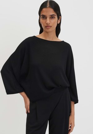 Vrouw met lang donker haar draagt een losse zwarte top met textuur en brede mouwen en zwarte broek, poserend tegen een effen lichte achtergrond.