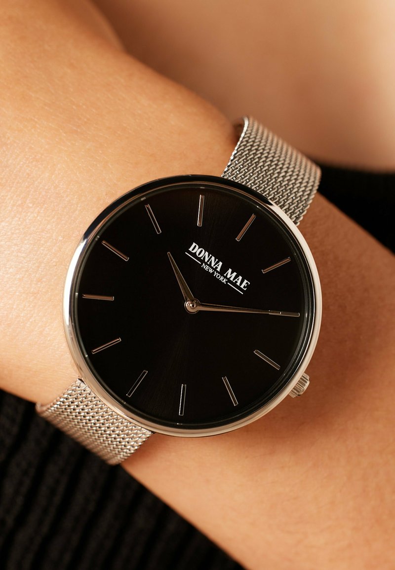 Zwarte wijzerplaat horloge met zilverkleurige mesh band, met minimalistische urenmarkeringen en merknaam "DONNA MAE" in witte tekst.