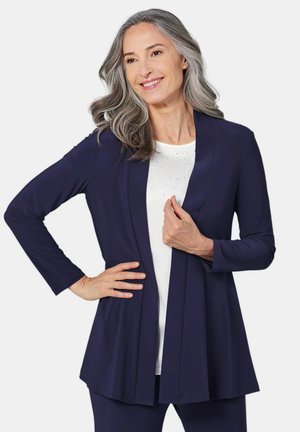Veste longue à manches longues de couleur marine avec une coupe décontractée, à l'avant ouvert et à la texture lisse, superposée à un haut blanc avec de petits accents brillants.