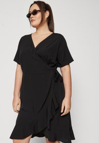 Crna haljina s omotom u mekom materijalu, s kratkim rukavima, vezanim strukom i volanima na dnu za dodatnu zanimljivost. Jednostavan i elegantan dizajn.