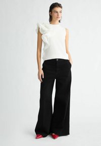 Top bianco senza maniche con dettaglio arricciato, abbinato a pantaloni neri a gamba larga e scarpe rosse con punta a stiletto. Colori solidi, texture eleganti.