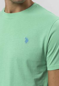 Mätová bavlnená tričko s okrúhlym výstrihom, so zelenomodrým vyšívaným logom na ľavej strane hrudníka. Mäkká textúra, krátke rukávy, klasický strih.