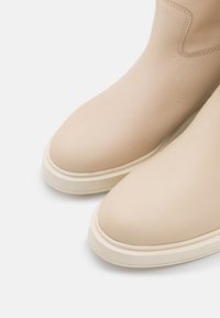 Pari nilon beji sepatu ankle dari kulit dengan ujung bulat dan sol karet putih tebal di latar belakang putih.