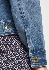 Veste en denim bleu clair avec un détail de bouton-pression doré sur le poignet, associée à un tissu noir à motifs, comportant de petites fleurs rouges et blanches.
