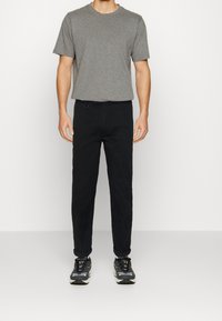 Graues kurzärmliges T-Shirt kombiniert mit schwarzen Hosen; das Modell trägt dunkle Turnschuhe. Das T-Shirt hat einen Rundhalsausschnitt, die Hose verfügt über Gürtelschlaufen.