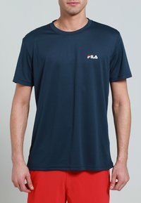 Muž nosící tmavě modré tričko s krátkým rukávem značky FILA a červené šortky, stojící před jednobarevným šedým pozadím.