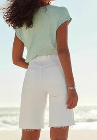 Une femme aux cheveux bouclés portant un t-shirt bleu clair et un short blanc effiloché se tient sur la plage, tournant le dos à l'océan sous un ciel dégagé.