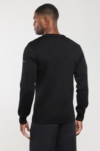 Hombre de espaldas con un suéter de manga larga negro y pantalones oscuros, de pie contra un fondo claro y liso.