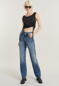 Crna skraćena majica bez rukava s nabranim stranama, uparena s svijetloplavim širokim trapericama i crnim gležnjačama. Model nosi sunčane naočale.