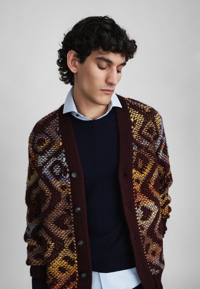 Knitted cardigan in maroon with geometric patterns in yellow, orange, and lavender; features buttons and a loose fit over a navy sweater and light shirt.