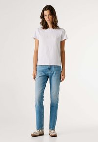 Sieviete ar plecu garumu matiem, ģērbusies baltā īsu piedurkņu krekliņā ar smalku tekstu, gaišbluejām džinsām un baltām rakstainām ikdienas apaviem, stāv.