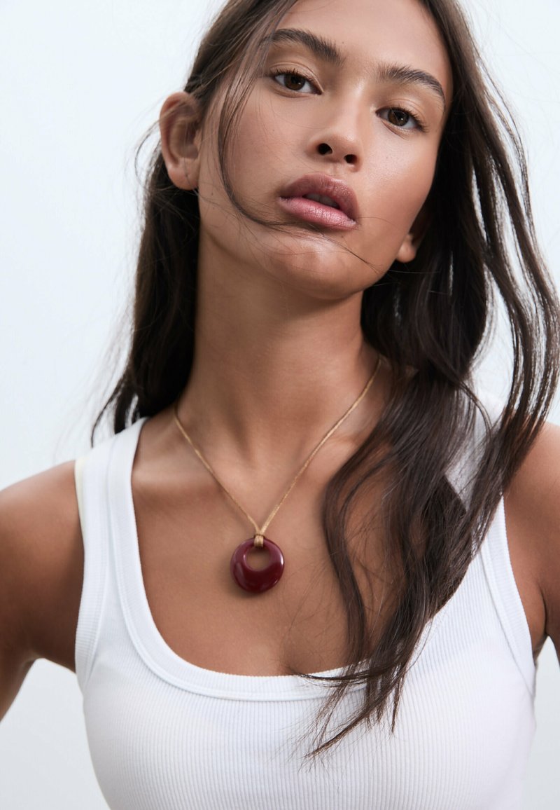 Collana d'oro con un pendente circolare di un profondo rosso, con un taglio centrale, indossata su una canottiera bianca a coste. Texture liscia e finitura lucida.