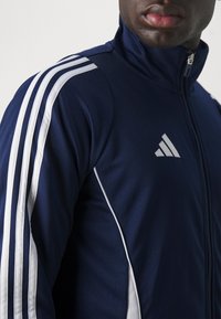 Marineblå jakke med glidelås og hvite striper på skuldrene, samt en liten sølvfarget logo på brystet. Glatt tekstur og atletisk design.
