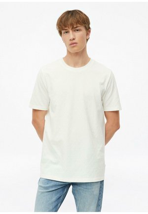 Młody mężczyzna o jasnobrązowych włosach, ubrany w zwykły biały T-shirt z krótkim rękawem i jasnoniebieskie jeansy, stoi z rękami za plecami.