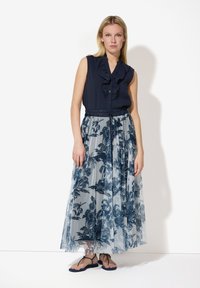 Blouse marine avec encolure volée, associée à une longue jupe transparente à motifs avec des designs floraux bleus et des sandales plates.