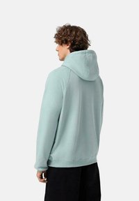 Sudadera verde menta con capucha ajustable, textura suave y puños acanalados, vista desde atrás, combinada con pantalones negros.