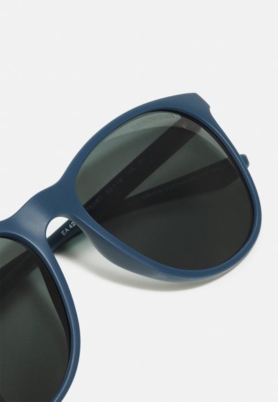 Occhiali da sole blu in plastica con finitura opaca e lenti scure. Presentano una montatura arrotondata e un dettaglio logo discreto sulla tempia.