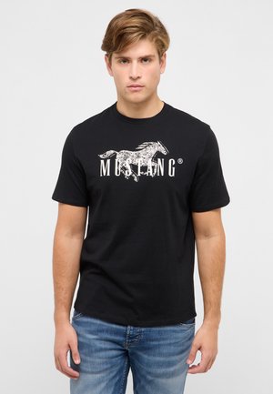 Junger Mann trägt ein schwarzes T-Shirt mit einem weißen, abgenutzten Grafikdruck eines galoppierenden Pferdes und dem Schriftzug "MUSTANG", kombiniert mit blauen Jeans, stehend vor einem einfarbigen Hintergrund.