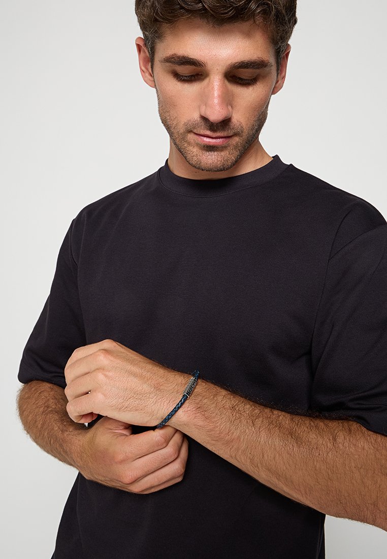 T-shirt noir à manches courtes porté par un mannequin, avec un bracelet bleu tressé orné d'accents métalliques au poignet.