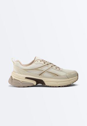 Beige low-top athletic sneaker with mesh and synthetic upper, cushioned sole, lace-up closure, and textured rubber outsole for traction.