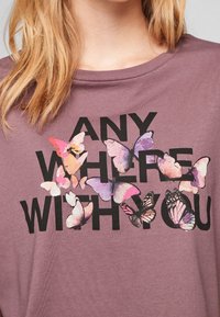 Fialové tričko s nápisem "KDEKOLI S TOBÍM" v tučných černých písmenech. Design obsahuje barevné grafiky motýlů rozptýlené po celém tričku.