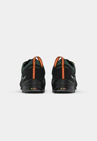 Sapatos atléticos pretos com um superior texturizado, detalhes verdes e uma etiqueta de puxar laranja brilhante no calcanhar; apresentando um design de sola de cor bloqueada.