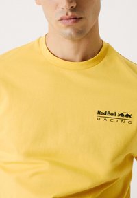 Gul bomuld t-shirt med rund hals, korte ærmer og et sort "Red Bull Racing" logo med en tilhørende grafik på venstre bryst.