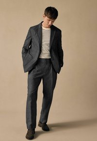 Traje gris a medida con solapas en forma de muesca y bolsillos frontales, combinado con una camisa a rayas. Los pantalones tienen un corte relajado y pliegue. Zapatos marrones.