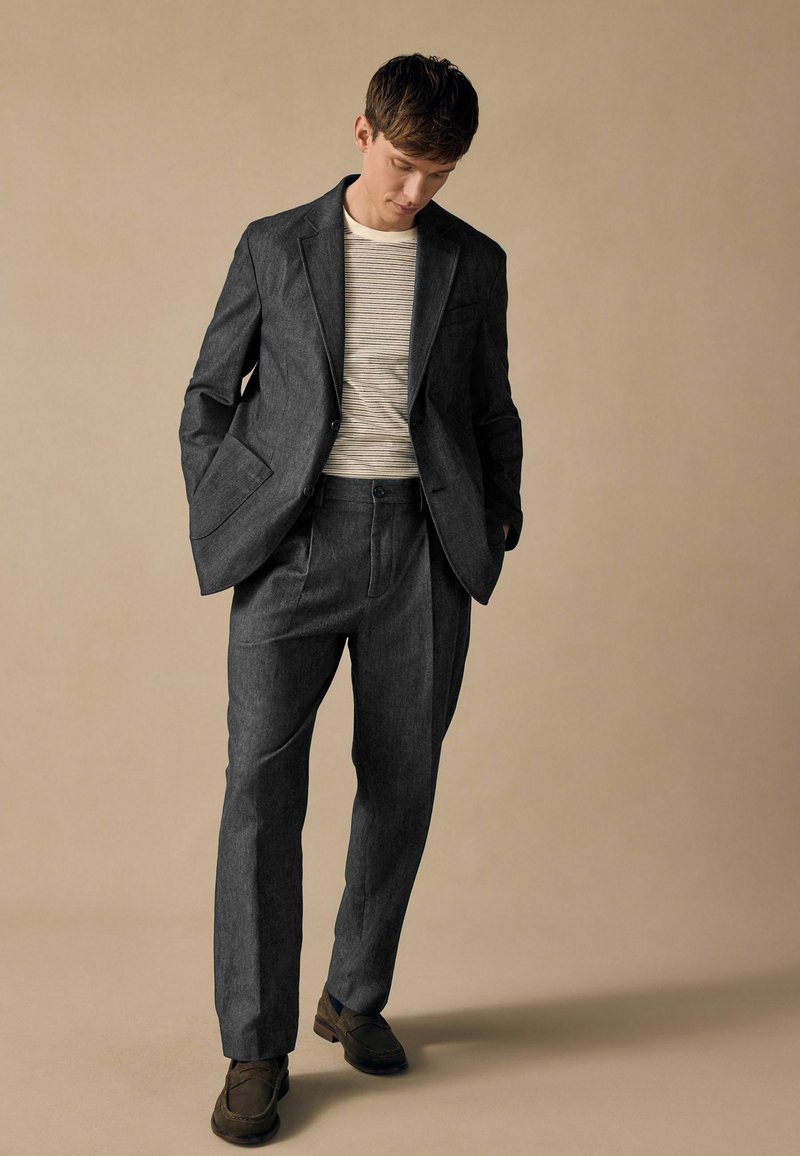 Traje gris a medida con solapas en forma de muesca y bolsillos frontales, combinado con una camisa a rayas. Los pantalones tienen un corte relajado y pliegue. Zapatos marrones.
