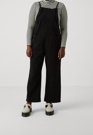 Salopette en denim noir à jambes larges et bretelles ajustables, associée à un col roulé rayé noir et blanc et des chaussures crème avec semelles noires.