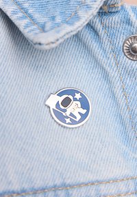 Srebrna emaliowana przypinka przedstawiająca astronautę na niebieskim okrągłym tle, akcentowana dwoma gwiazdkami, przymocowana do jasnoniebieskiego denimu.