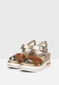 Sandalias cuña con correas de cuero marrón y dorado, un hebilla de tobillo segura y suela de goma blanca con una franja de acento dorado.