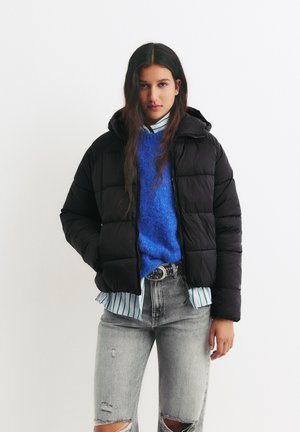 Zwarte pufferjack met capuchon, gewatteerd design, gedragen over een blauwe trui en een gestreepte shirt, gecombineerd met versleten lichtgrijze jeans.