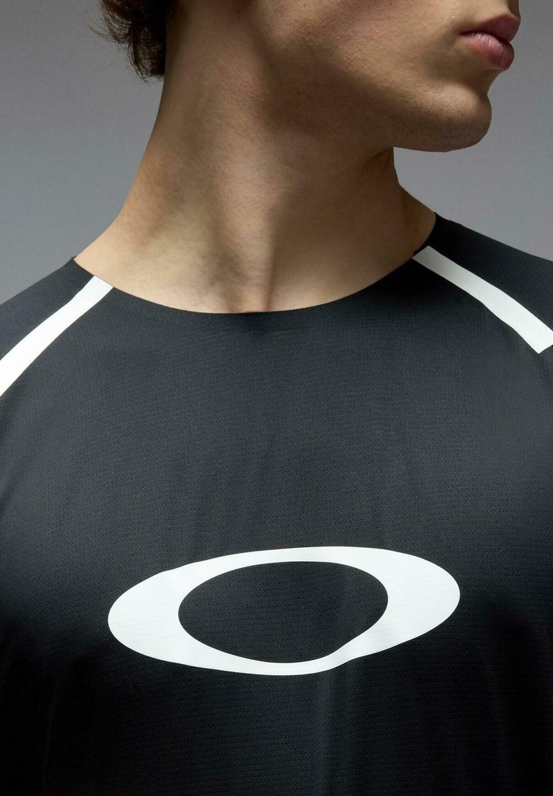 Jeune personne portant un maillot de sport noir avec un grand logo ovale blanc sur la poitrine, vue de profil sur un fond uni.