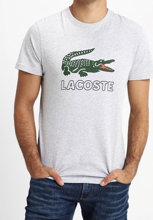 Szara bawełniana koszulka z zielonym logo krokodyla i czarnym napisem "LACOSTE". Prosty, okrągły dekolt, krótkie rękawy.