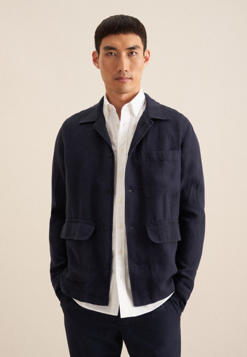 Giacca di lino blu navy con colletto, chiusura frontale con bottoni, due tasche a toppa e maniche lunghe, indossata sopra una camicia bianca con bottoni.