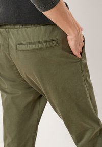 Persoon in olijfgroene broek met een paspel achterzak, hand in zijzak, en een donkergrijze top zichtbaar op taillehoogte.