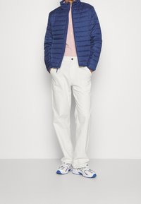 Marineblauwe donsjas met horizontale stiksels, lichtroze blouse met knopen, witte broek met wijde pijpen, en grijsblauwe sneakers met blauwe accenten.