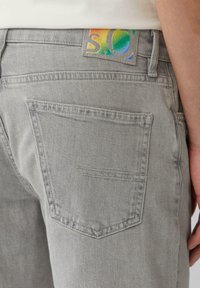 Jean en denim gris avec une poche arrière et une étiquette en tissu colorée avec le logo "S.Q". Texture lisse avec un design classique à cinq poches.